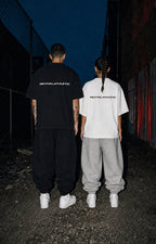 Two models from behind wearing oversized REVIVALLATHLETIC t-shirts in black and white, paired with baggy sweatpants and white sneakers, standing in a dark alley at night.
