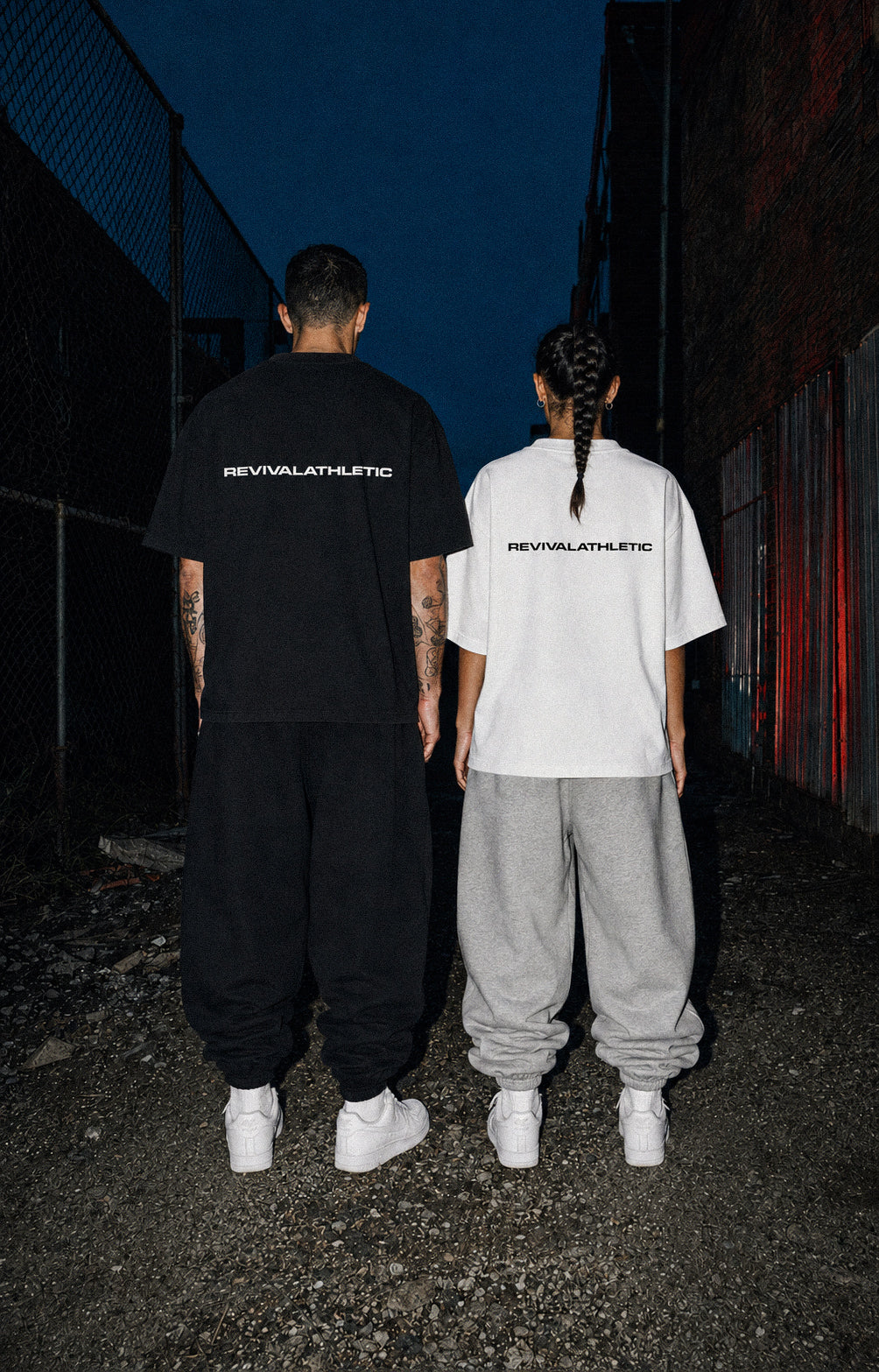 Two models from behind wearing oversized REVIVALLATHLETIC t-shirts in black and white, paired with baggy sweatpants and white sneakers, standing in a dark alley at night.