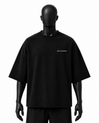 A black oversized short-sleeve t-shirt displayed on a matte black mannequin against a plain white background. The shirt features a small white script logo on the left chest.