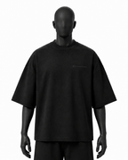 A black oversized short-sleeve t-shirt with a small tonal script logo on the left chest, displayed on a matte black mannequin against a plain white background.